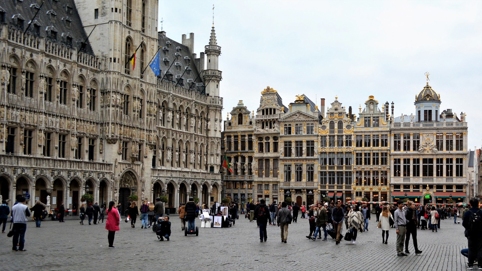 The Grand Place in Brussels, with the Gothic spire of the Town Hall on the left and gilded 17th-century guild houses on the right, busy with tourists crossing the cobblestone square.