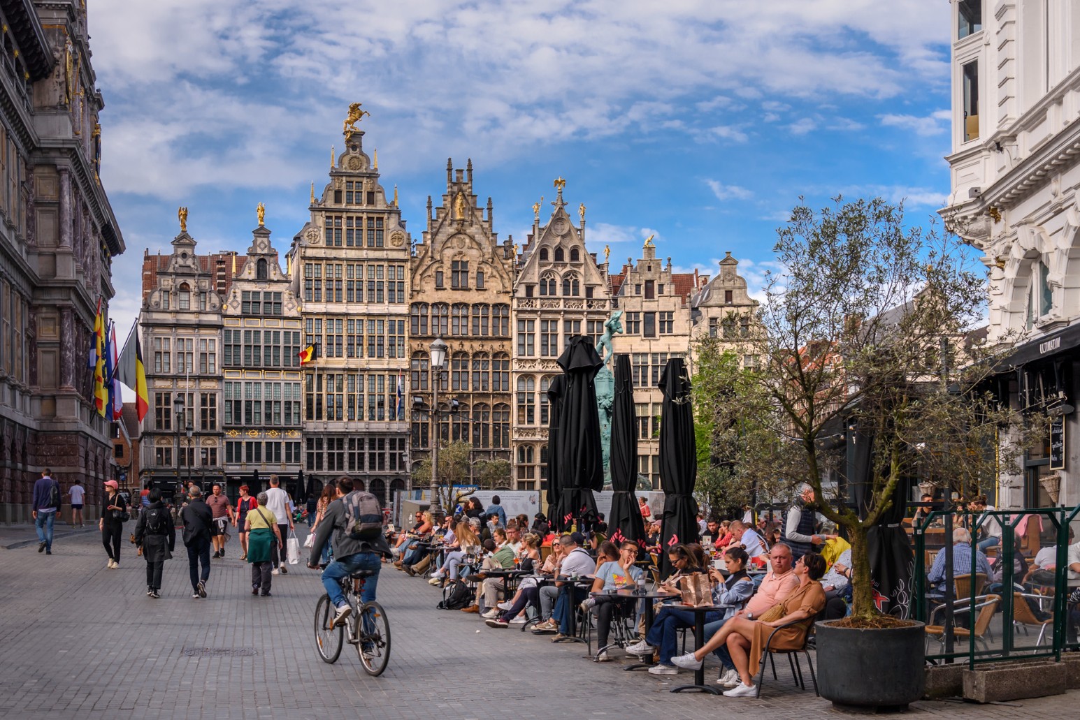 Antwerp's Grote Markt lined with ornate gabled guild houses, with outdoor café tables, a cyclist passing through, and the Brabo fountain statue in the square.