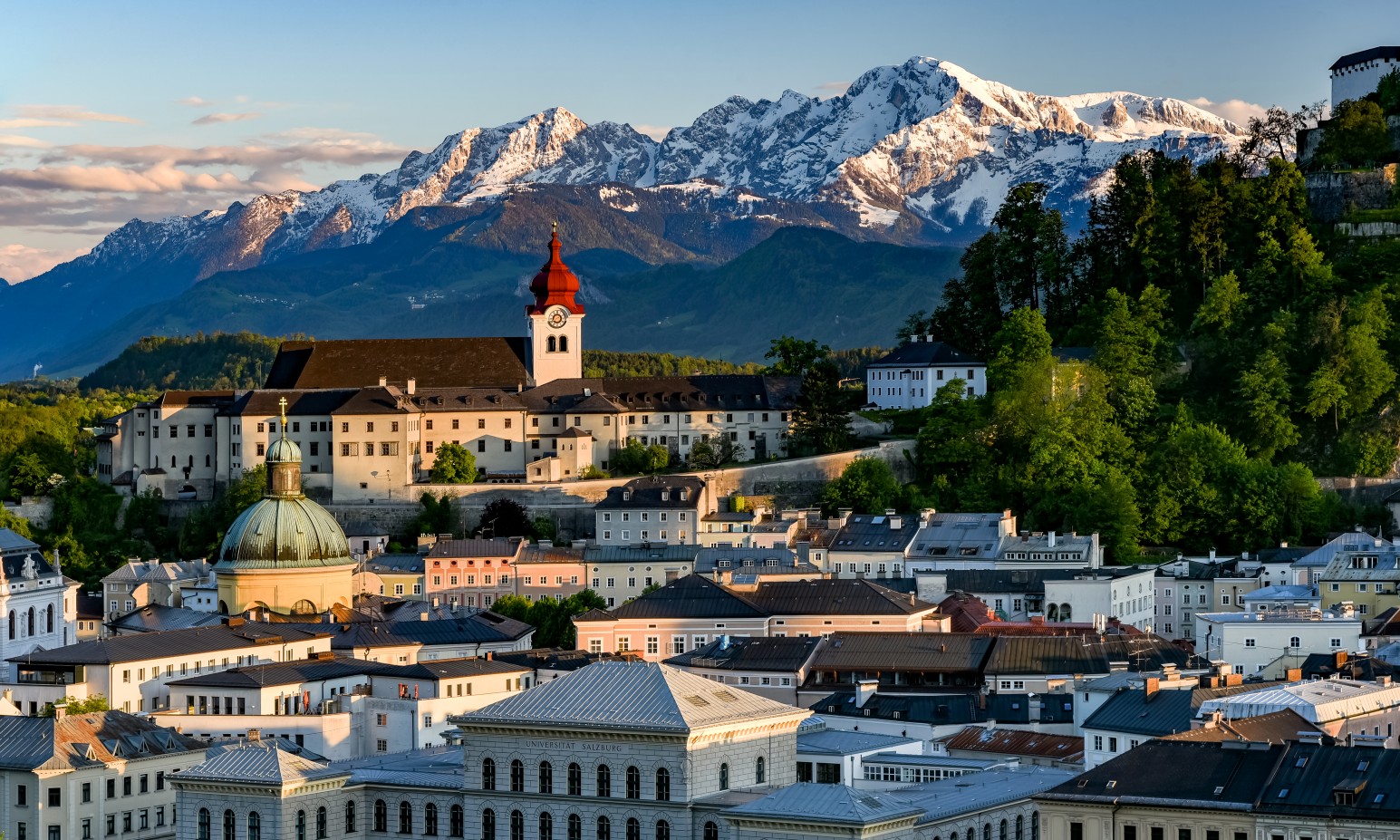 The rooftops and church towers of Salzburg's old town at sunset, with the snow-capped Alps rising behind the city.
