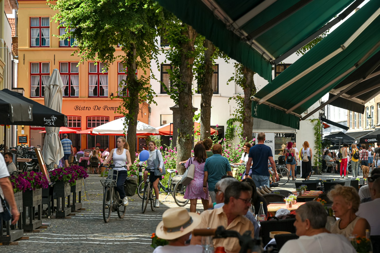 A tree-lined cobblestone street in Bruges with outdoor café seating, cyclists, and colourful gabled buildings including the Bistro De Pomp restaurant.