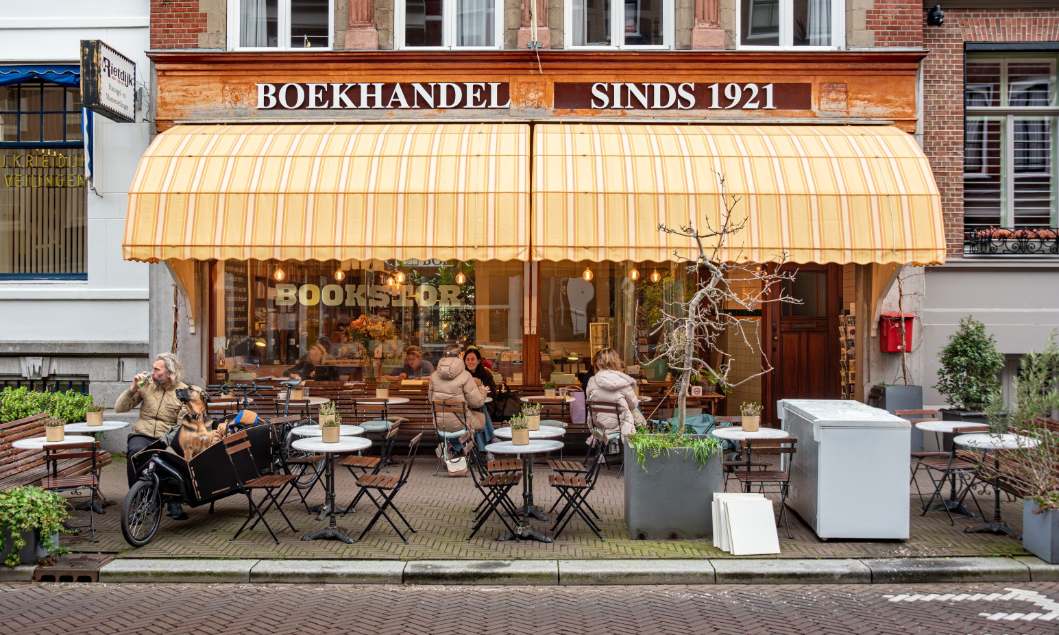 A bookshop terrace in the Netherlands under a striped awning reading 'Boekhandel Sinds 1921,' with people seated at outdoor café tables and a cargo bike carrying a dog parked alongside.