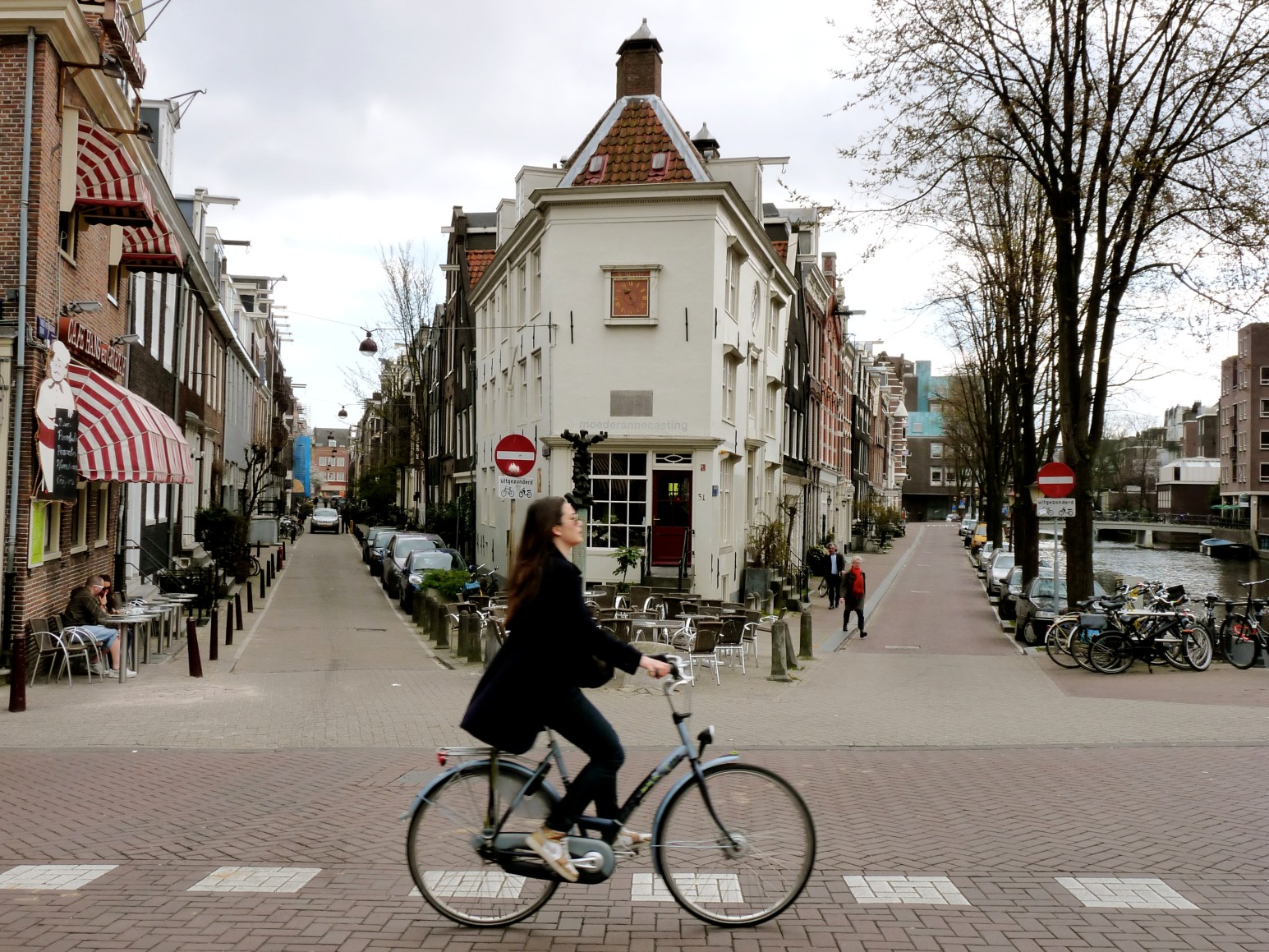 A cyclist crossing a brick street in Amsterdam past a distinctive triangular gabled building with a clock, canal houses, and bicycles lined up along a canal.