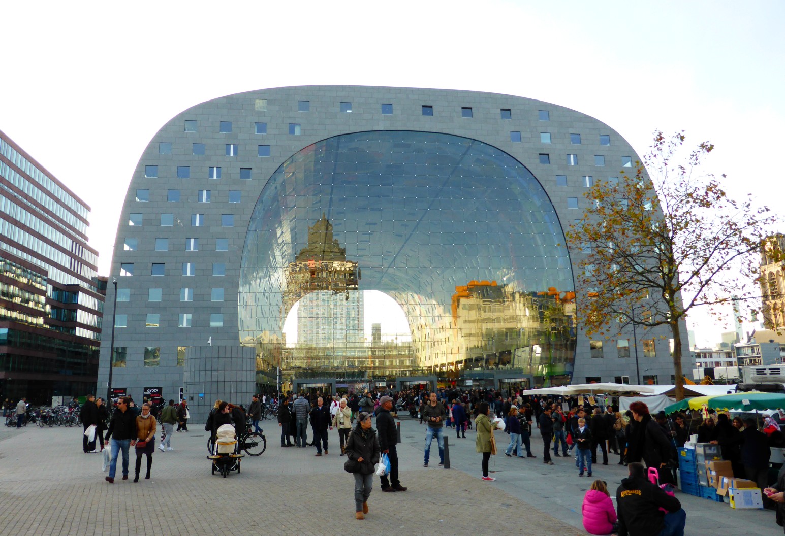 The Markthal in Rotterdam, a large arched building with a glass window front reflecting the surrounding cityscape, with crowds and market stalls in the square below.