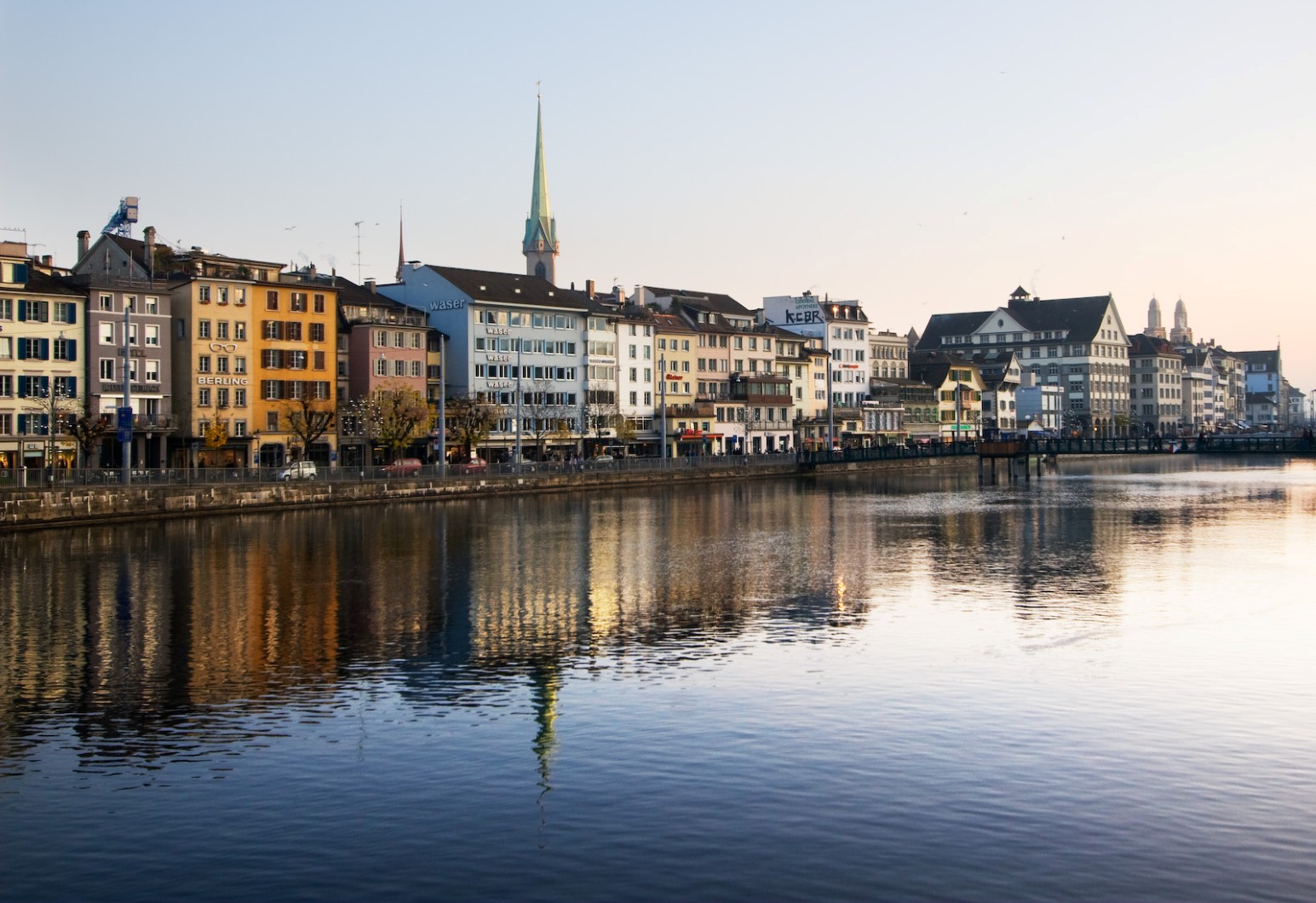 The Limmat river in Zurich at dusk, lined with colourful historic buildings and a church spire, all reflected in the calm water.