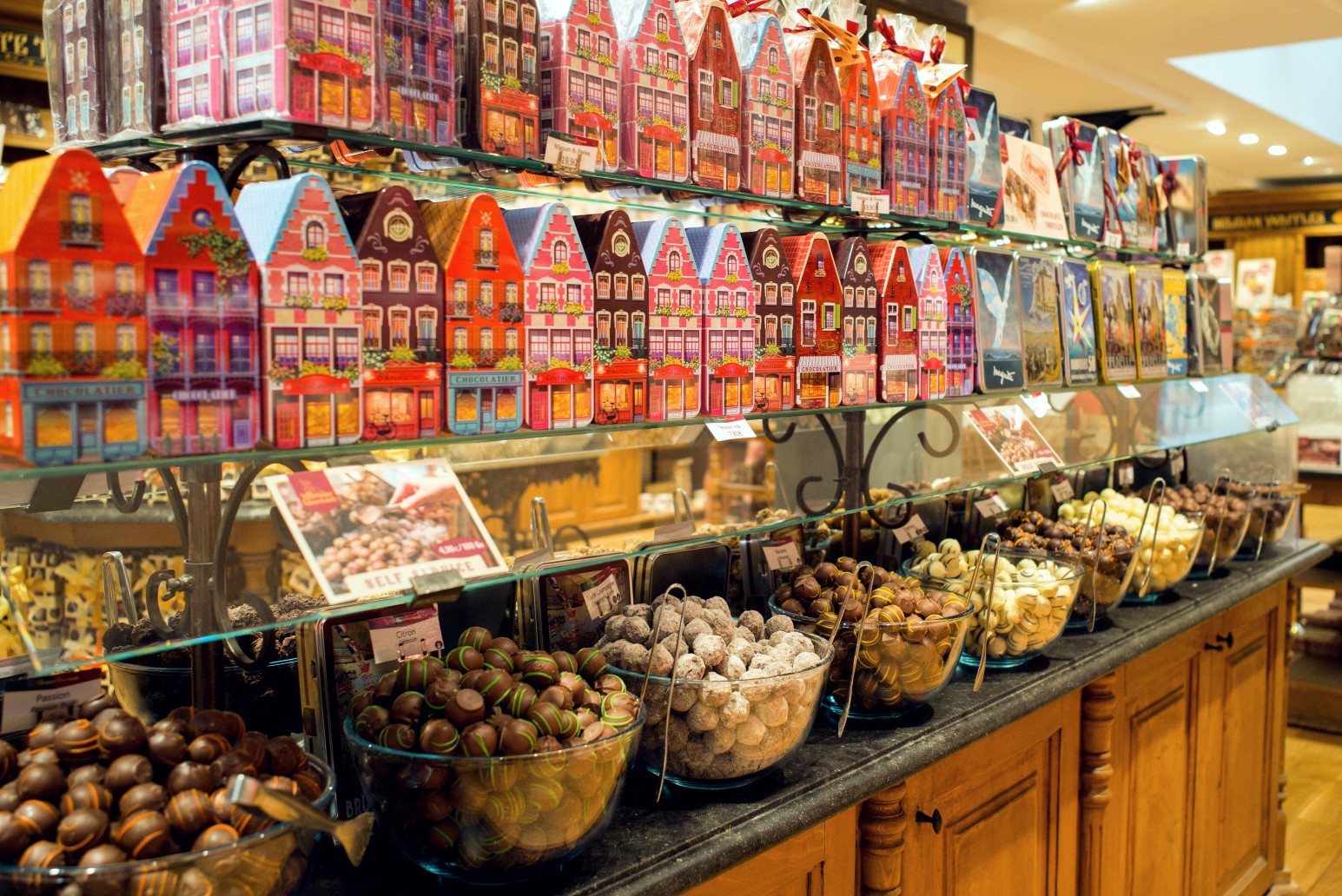 A Belgian chocolate shop display, with rows of colourful gabled-house-shaped gift tins on glass shelves above bowls of assorted chocolate truffles and pralines.