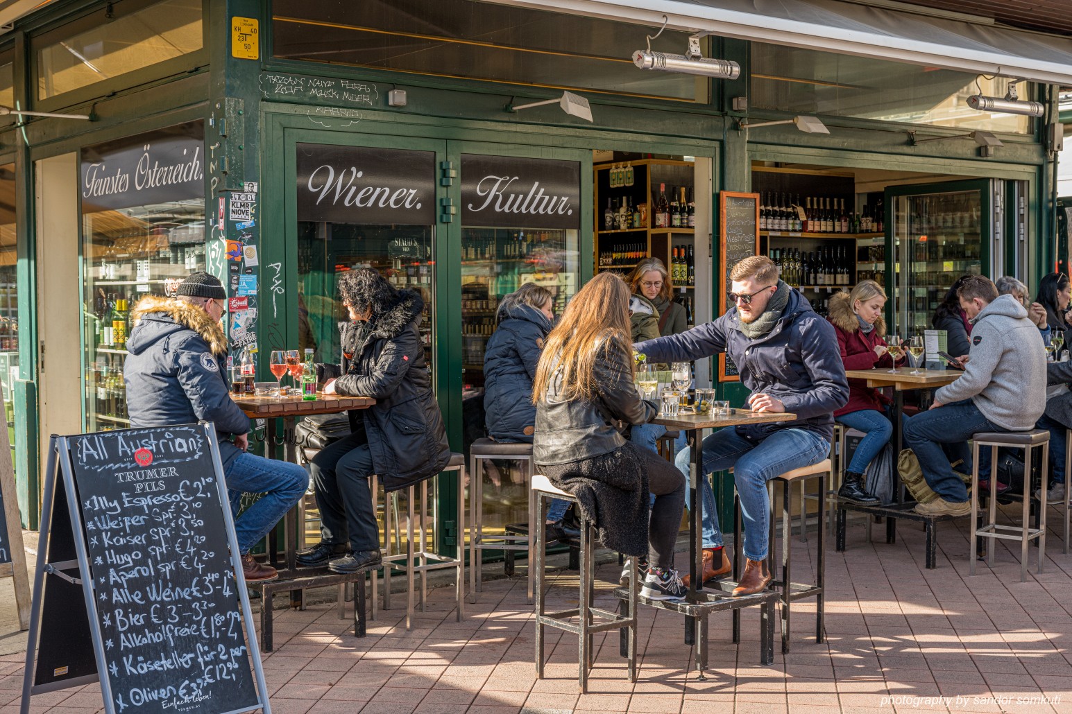 Diners sitting at outdoor high tables outside a Vienna wine bar with "Wiener Kultur" signage and a chalkboard drinks menu.