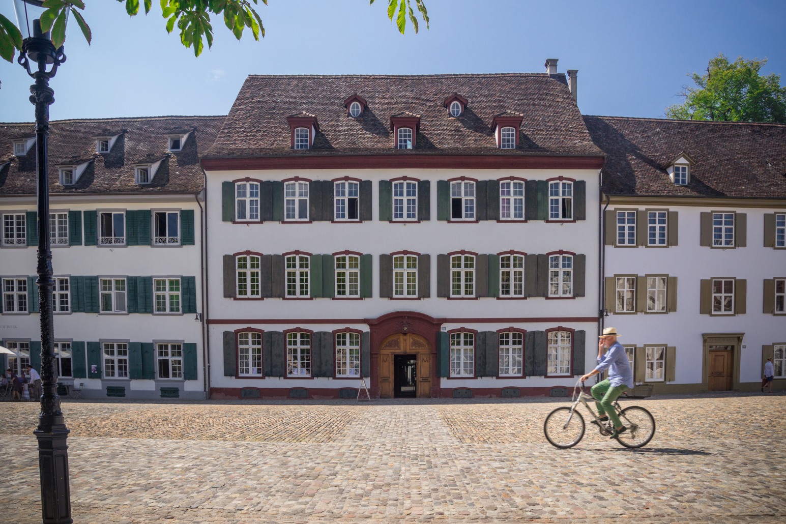 A grand red-and-white shuttered building on a cobblestone square in Basel, with a cyclist riding past in the foreground.