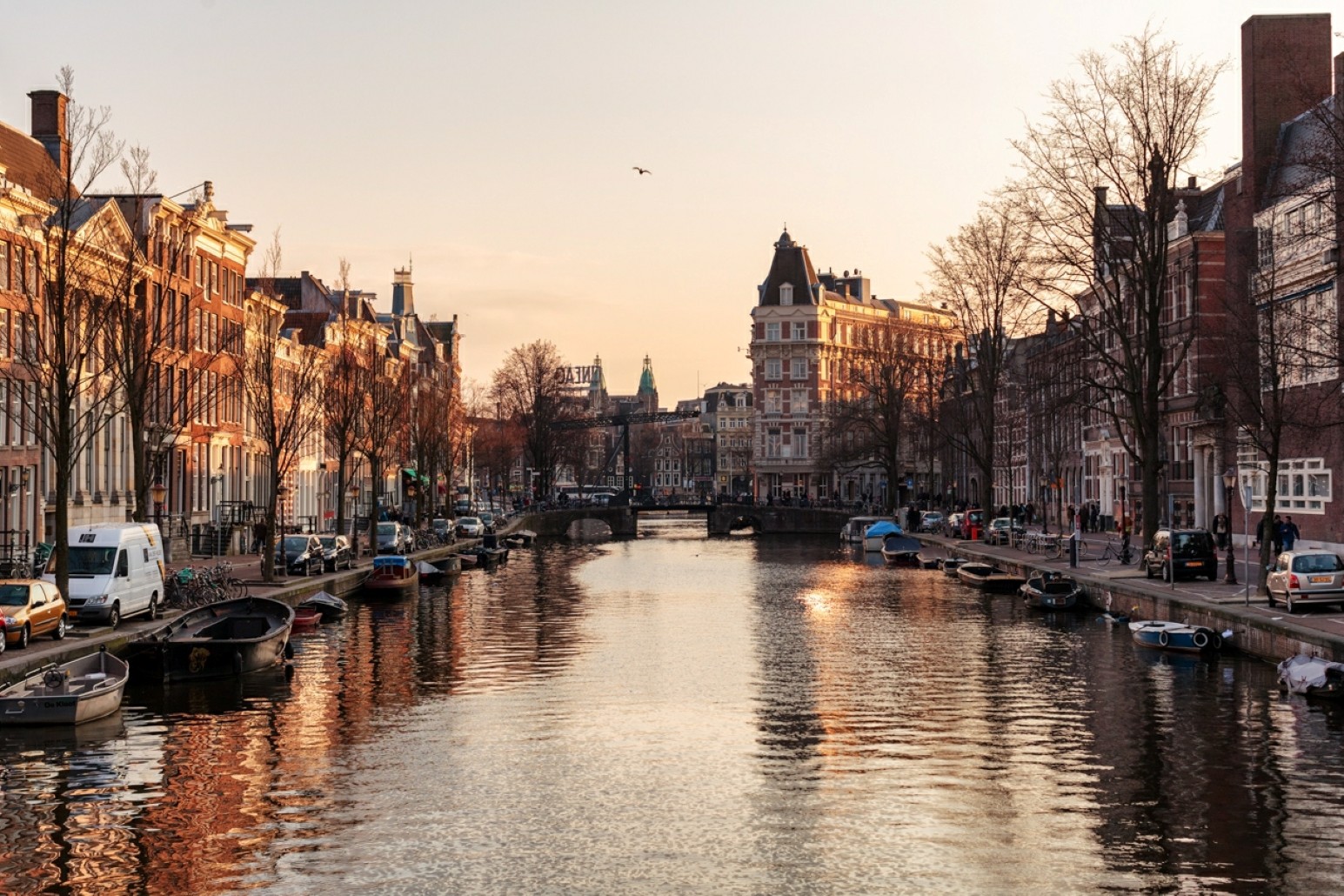 An Amsterdam canal at sunset lined with historic gabled townhouses, small boats moored along the water, and a bridge in the distance.
