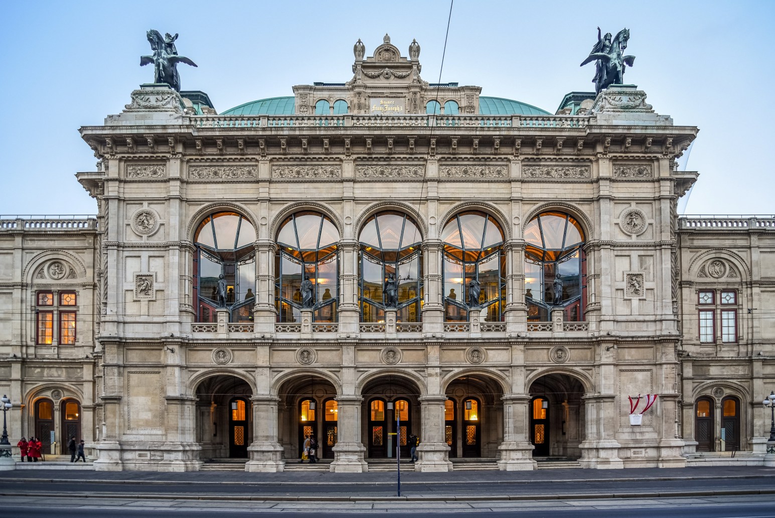 The ornate facade of the Vienna State Opera House at dusk, with bronze horse statues on the roofline and glowing arched windows along its balcony.