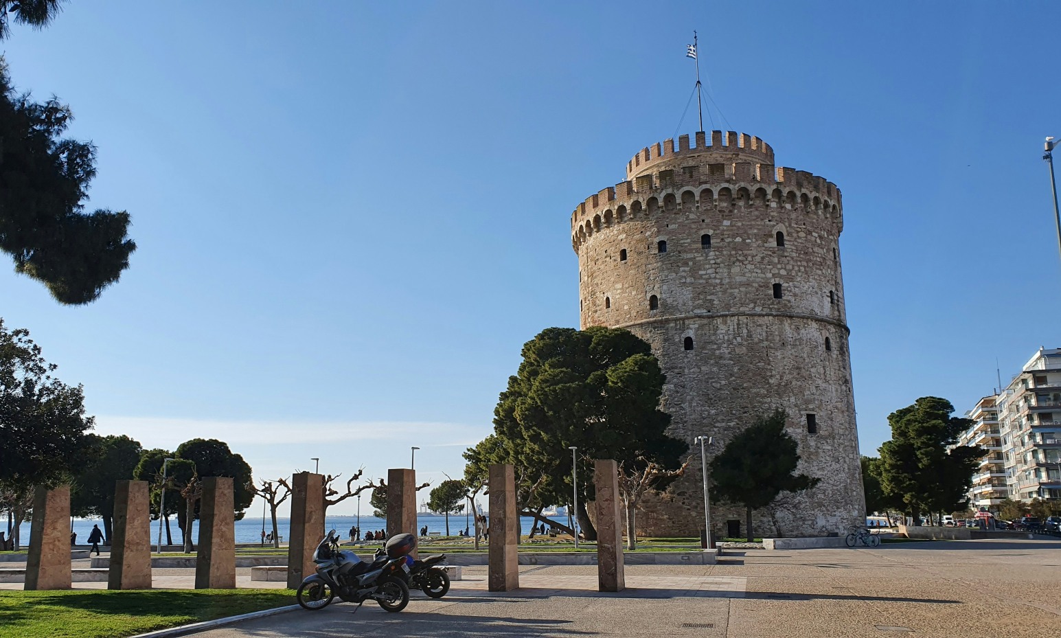 The White Tower of Thessaloniki, a cylindrical stone fortress with battlements on the waterfront promenade, surrounded by trees and stone memorial pillars.