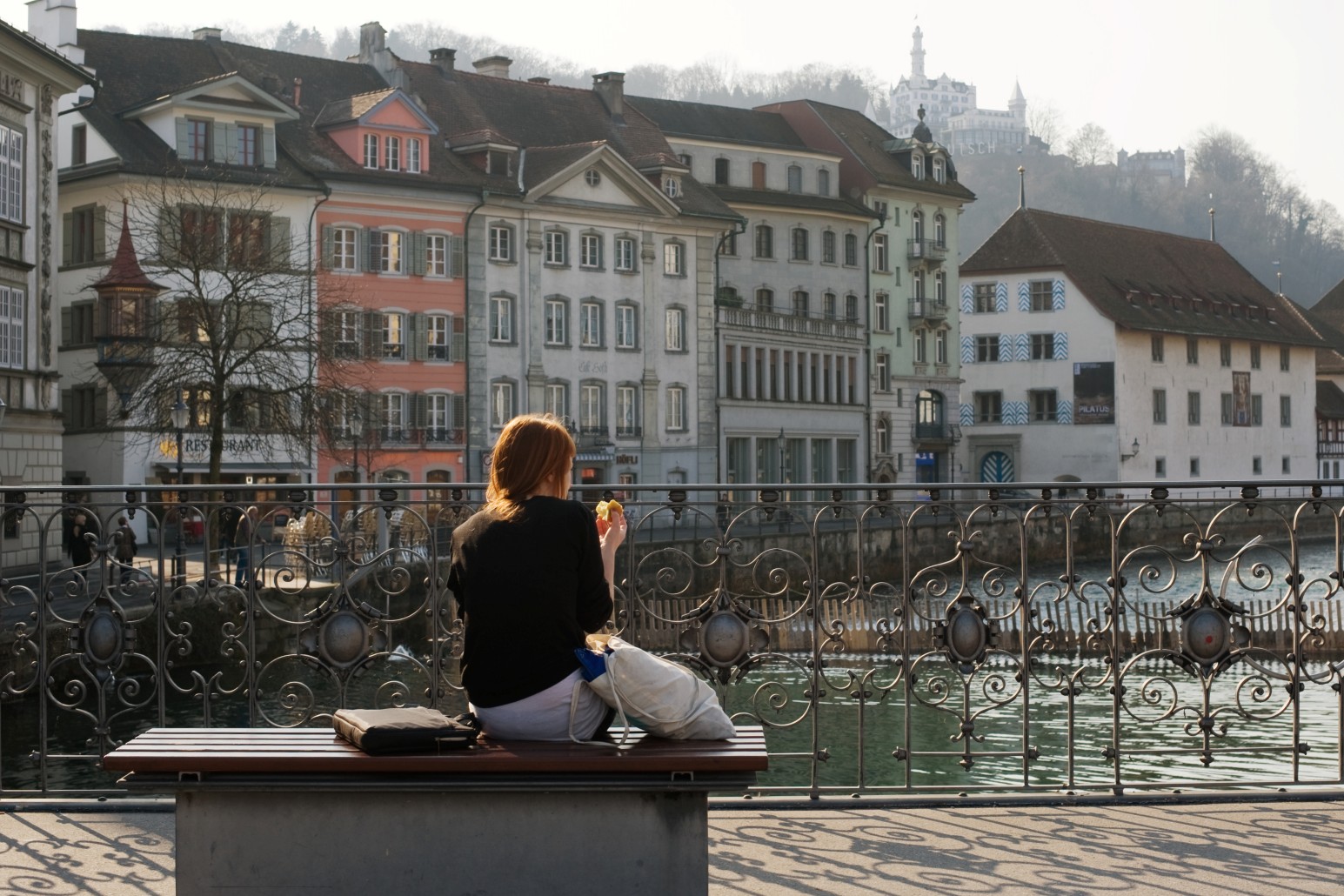 A woman sitting on a riverside bench in Lucerne, eating a pastry and looking out over an ornate iron railing at colourful historic buildings along the water.