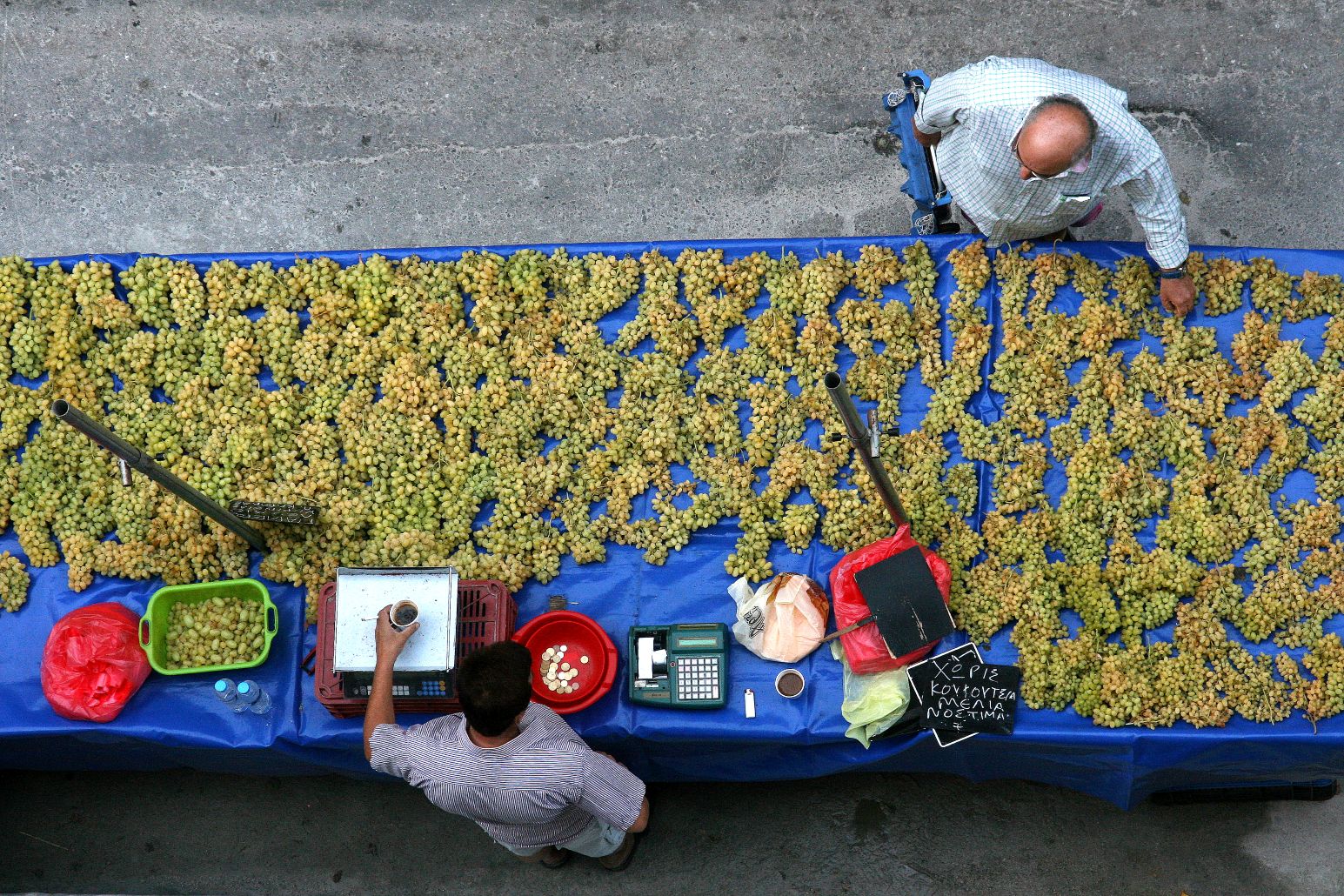 An overhead view of a street vendor's stall in Athens with bunches of fresh grapes laid out on a blue tarp, alongside scales, a cash register, and a handwritten Greek price sign.