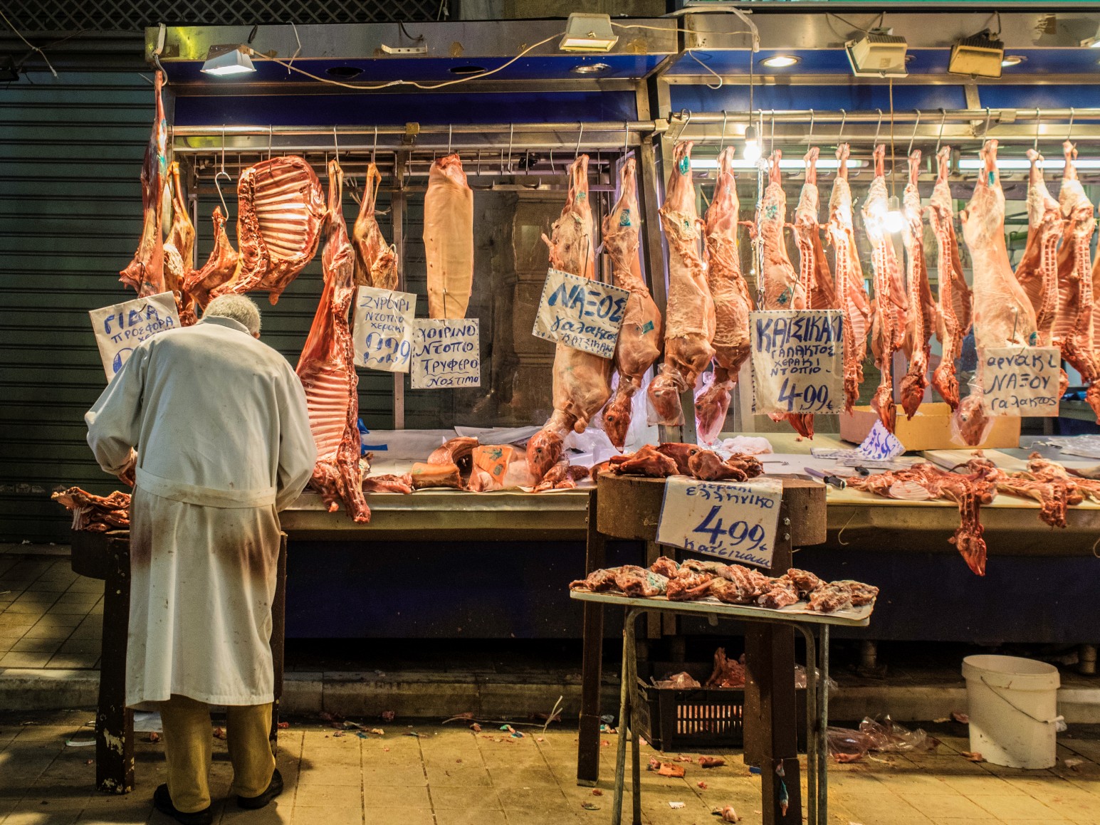 A butcher's stall at a Greek meat market with whole carcasses hanging on display, handwritten price signs in Greek, and a butcher in a white coat serving a customer.