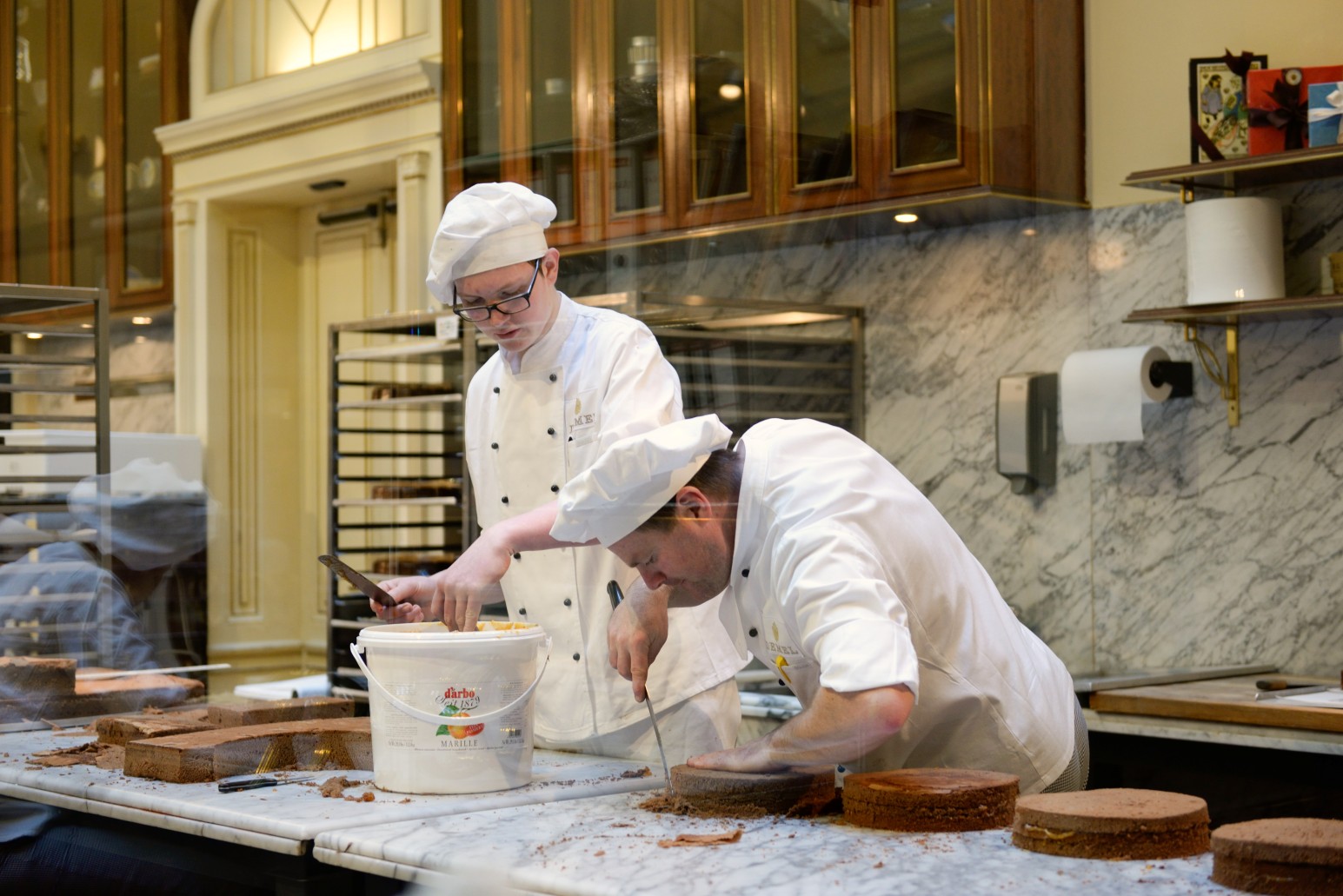 Two chefs in white uniforms and toques carefully layering and trimming chocolate sponge cakes at a marble counter in a bakery kitchen.