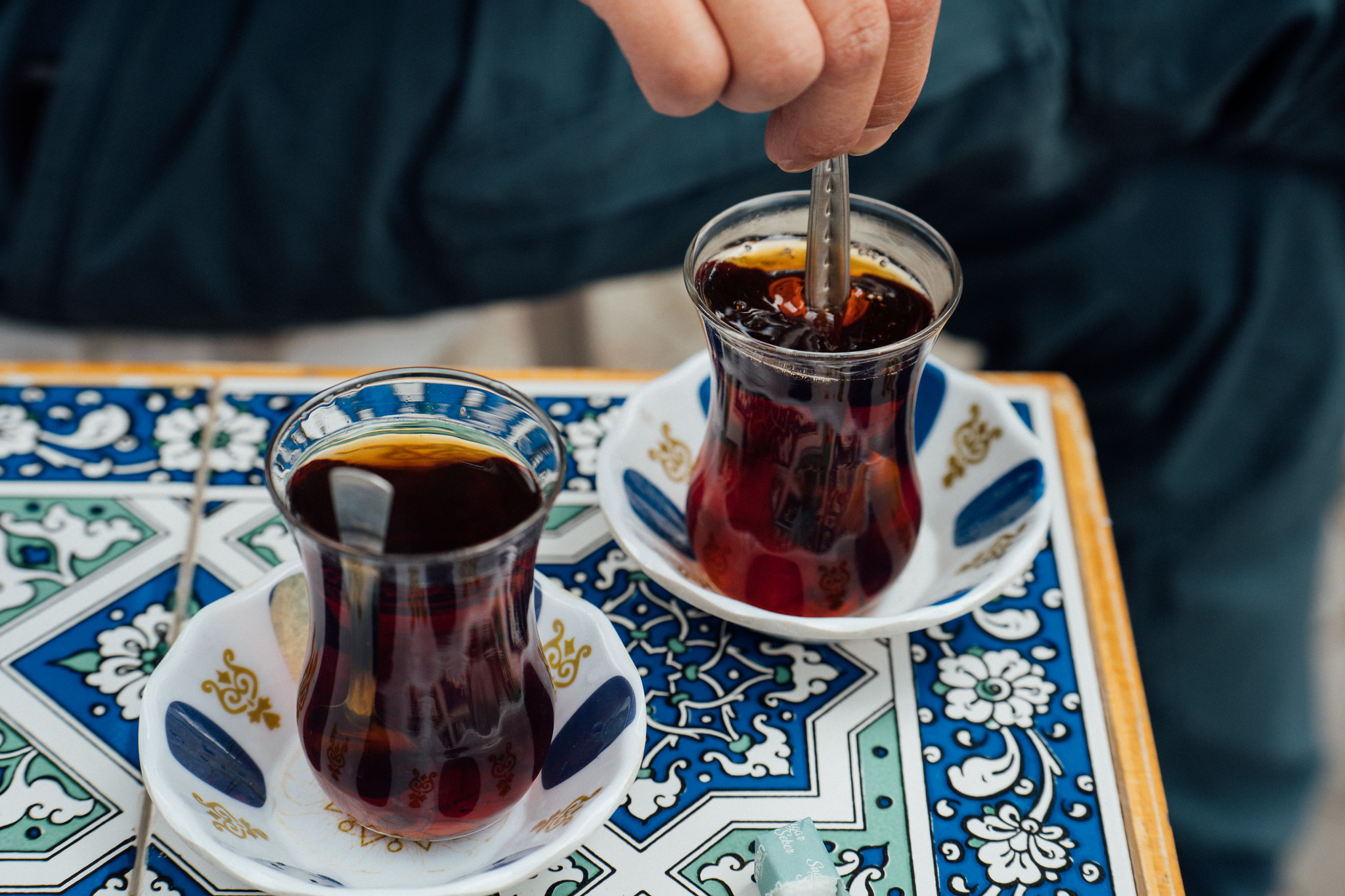 Two glasses of Turkish tea on tulip-shaped saucers atop a blue and white patterned table, one being stirred