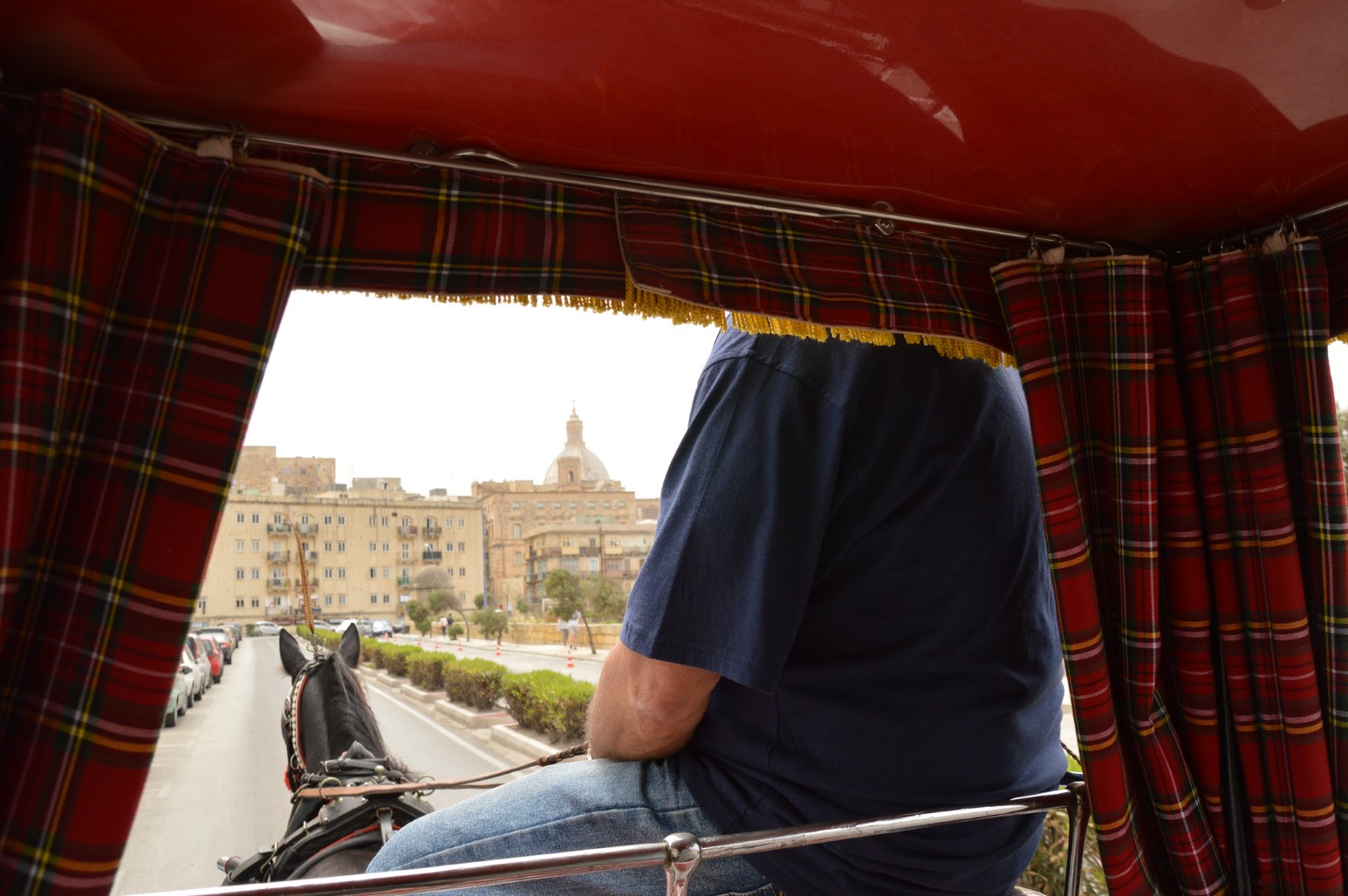 A view from inside a traditional horse-drawn karrozzin carriage looking out over a tasselled tartan curtain onto a Valletta street and domed church.