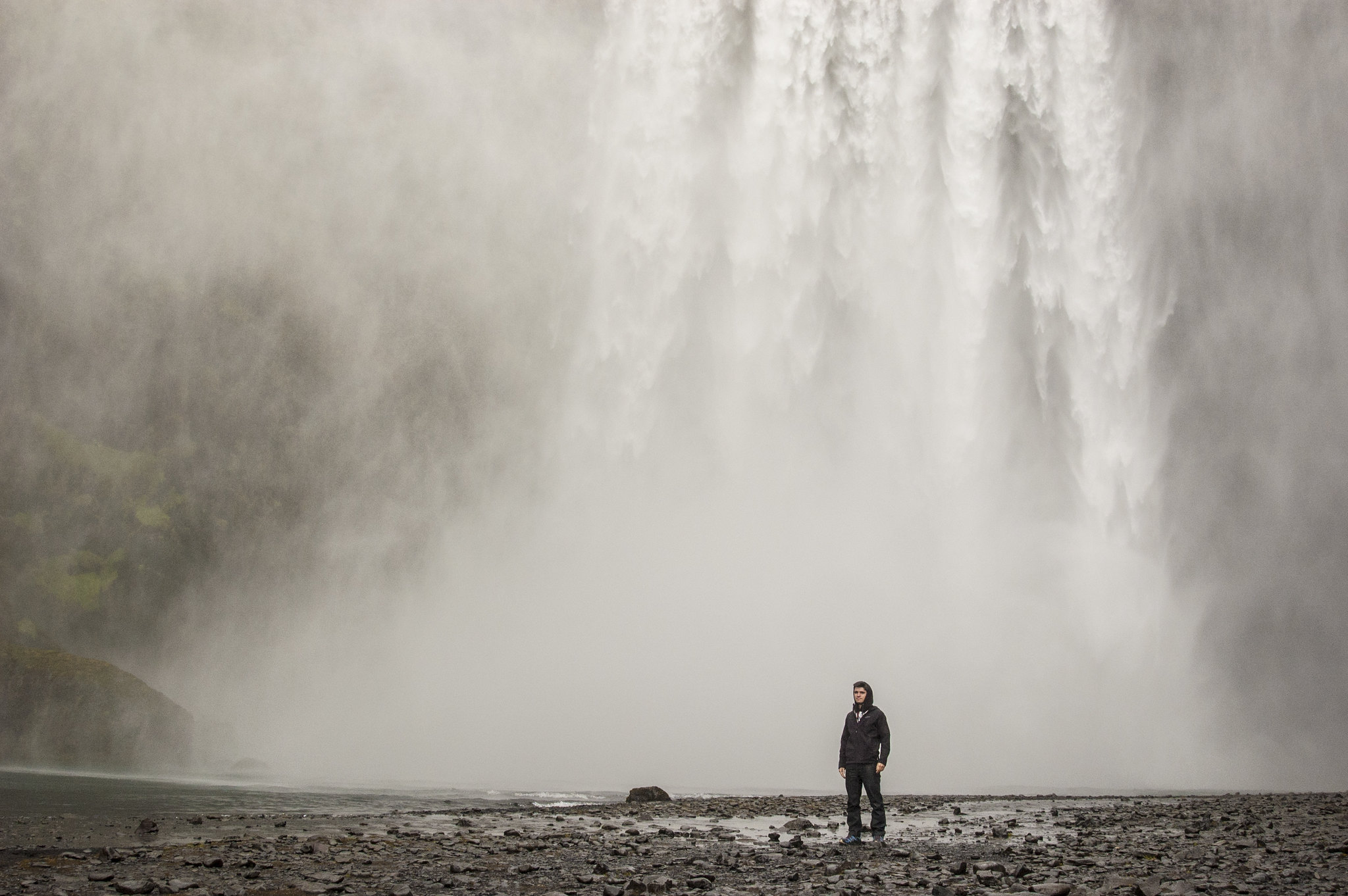 A lone figure standing on a black rock beach beneath the thundering spray of Skógafoss waterfall