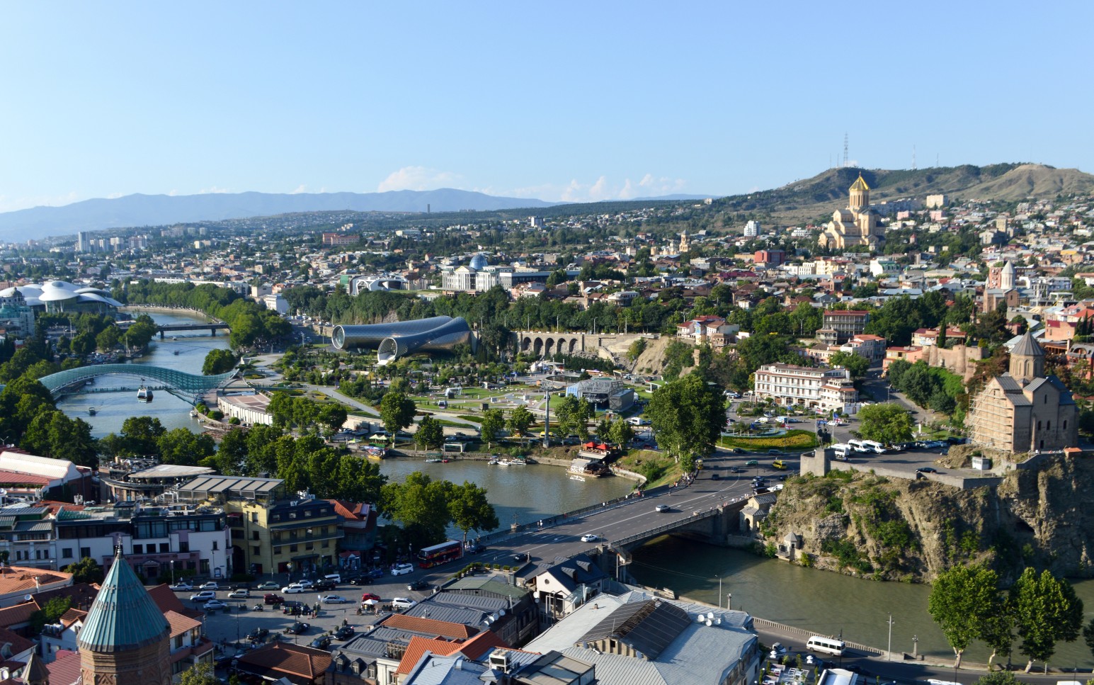 Aerial view of Tbilisi with the Bridge of Peace crossing the Mtkvari River, Rike Park's tubular concert hall, and the Sameba Cathedral on a hilltop in the distance