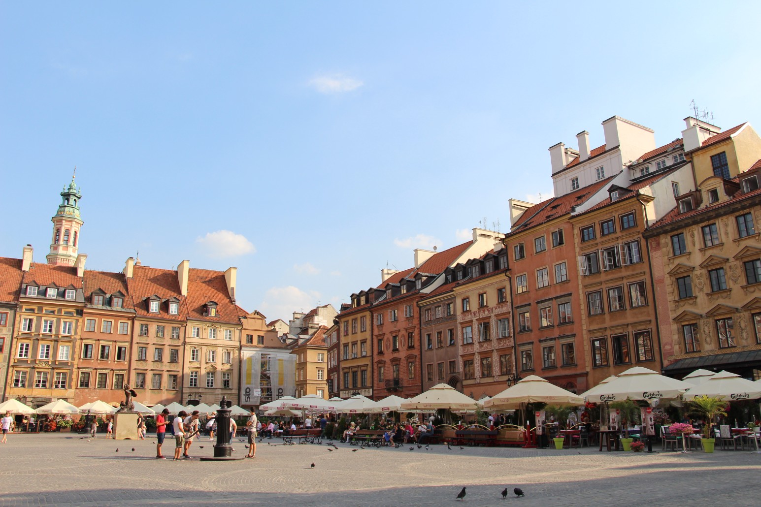 The colourful historic townhouses of Warsaw's Old Town Market Square, with outdoor café umbrellas and pigeons gathered in the open square below.
