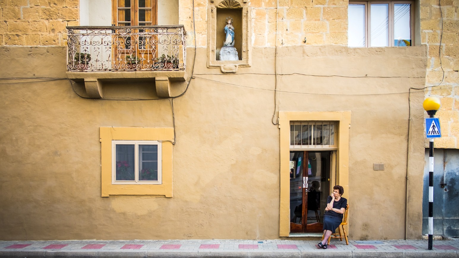 An elderly woman sitting in a doorway chair outside a sandstone house in Kercem, beneath a wrought-iron balcony and a small statue niche.