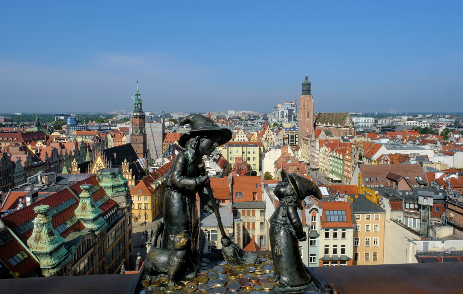 A rooftop view over Wrocław's colourful old town, with bronze witch statues in the foreground and church spires rising above the rooftops.