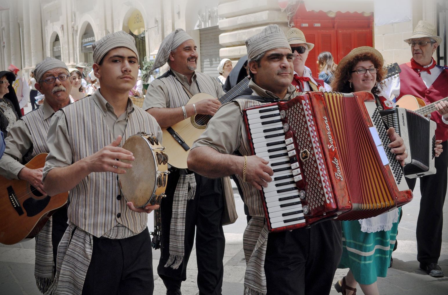 A traditional Maltese folk band in striped headscarves playing accordion, guitar, and tambourine during a street festival procession.