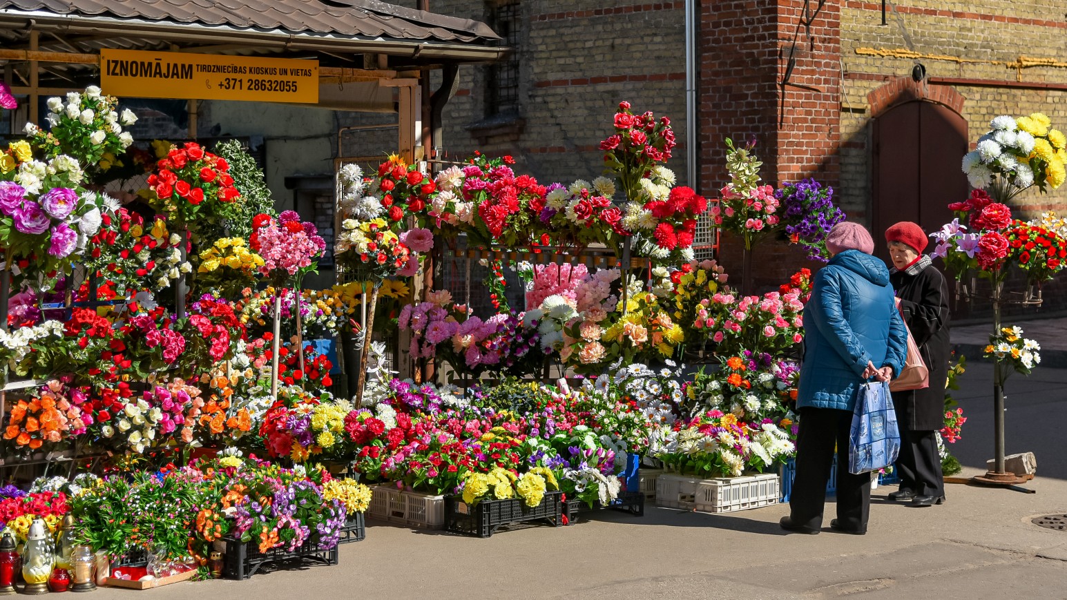 Two women browsing a colourful outdoor flower stall piled with bouquets outside Riga Central Market.