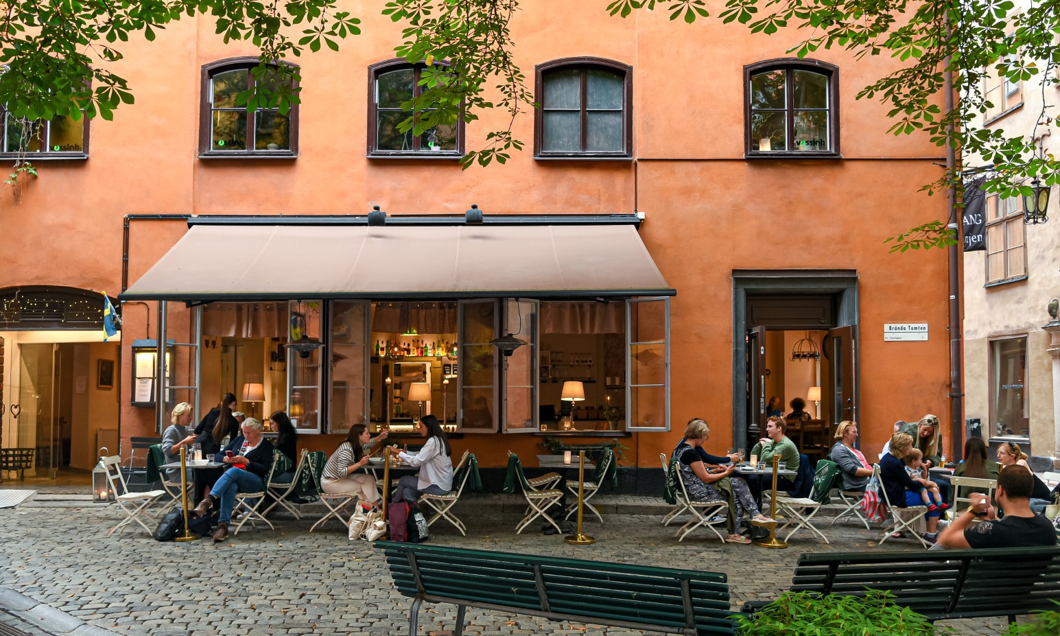 People sitting at outdoor café tables outside an orange building on a cobblestone square in Stockholm, Sweden