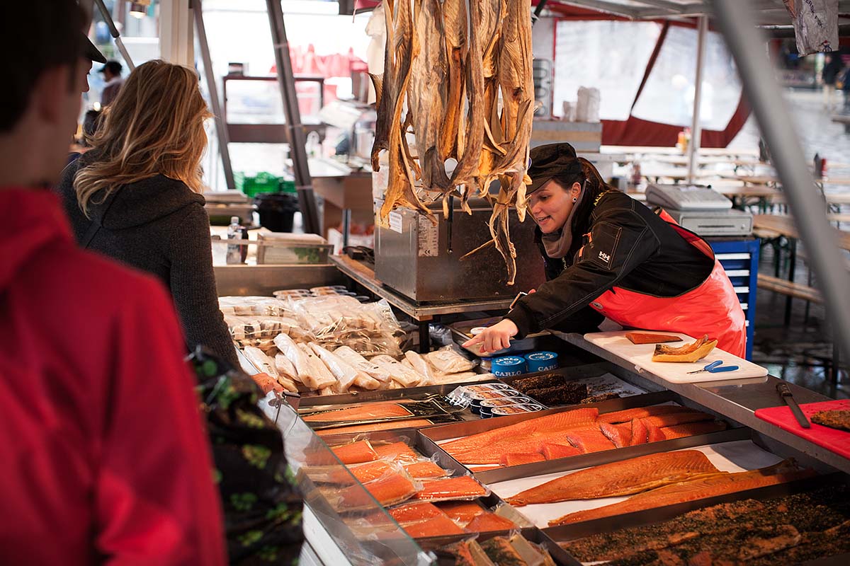 A fishmonger serving customers at an outdoor seafood stall with dried fish hanging overhead and fresh salmon on display, at a market in Bergen, Norway