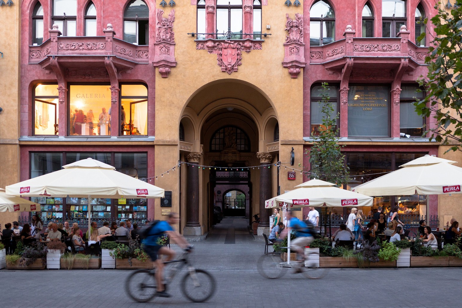 An ornate pink and yellow Art Nouveau building in Łódź with an arched entrance, outdoor café seating, and cyclists passing by at dusk.
