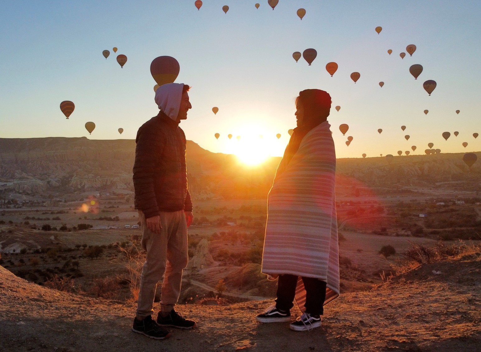 Couple wrapped in a blanket watching hot air balloons fill the sky at sunrise over Cappadocia's valleys