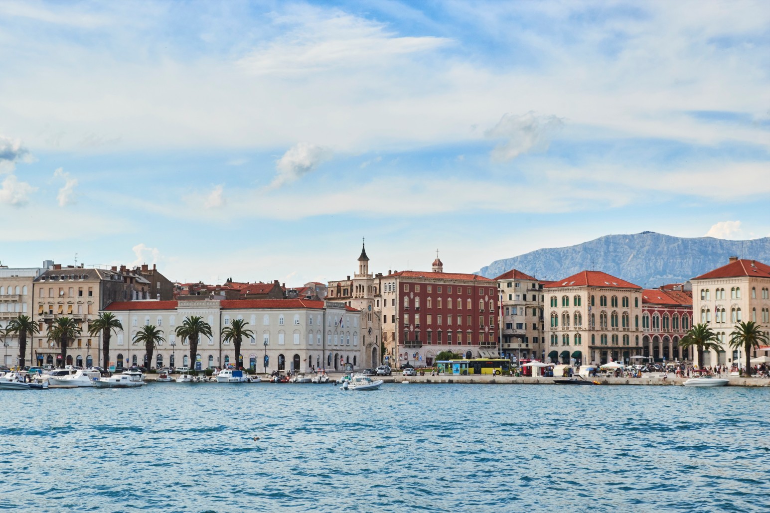The waterfront promenade of Split, Croatia, lined with palm trees, historic buildings, and moored boats, with mountains visible across the bay.