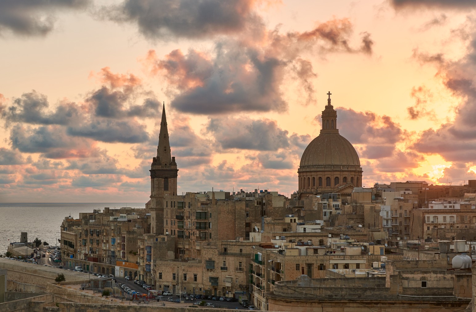 The sandstone skyline of Valletta at sunset, with the dome of the Carmelite Church and a bell tower silhouetted against a dramatic cloudy sky.