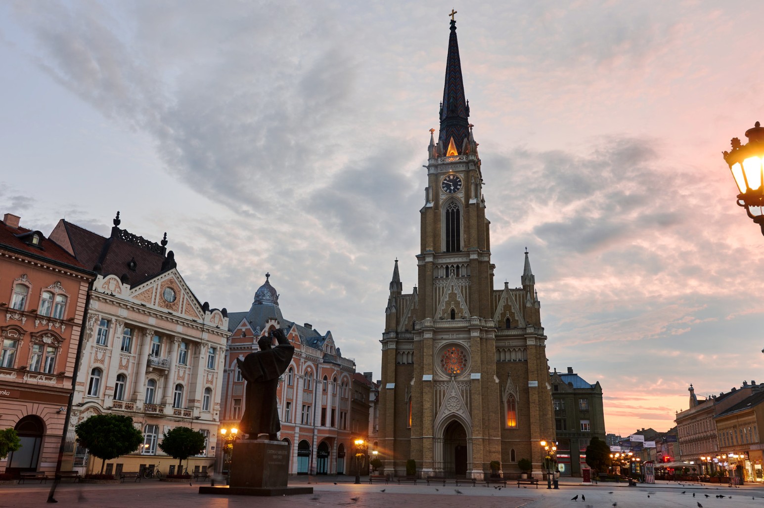 Name of Mary Church in Novi Sad’s main square at sunset, with a statue in the foreground and pastel historic buildings alongside
