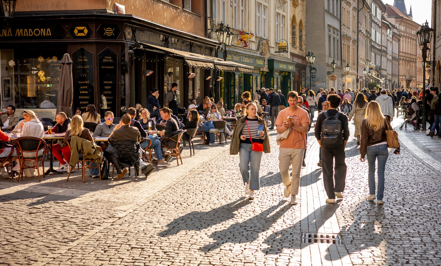 Crowds of tourists and diners walking and sitting at outdoor cafés along a sunlit cobbled street in Prague's old town.