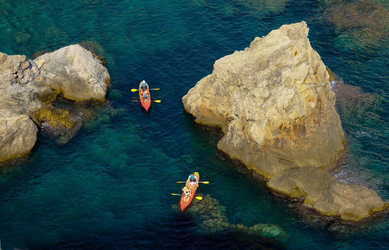 An aerial view of two kayaks paddling through turquoise water between large rocky outcrops on the Croatian coast.