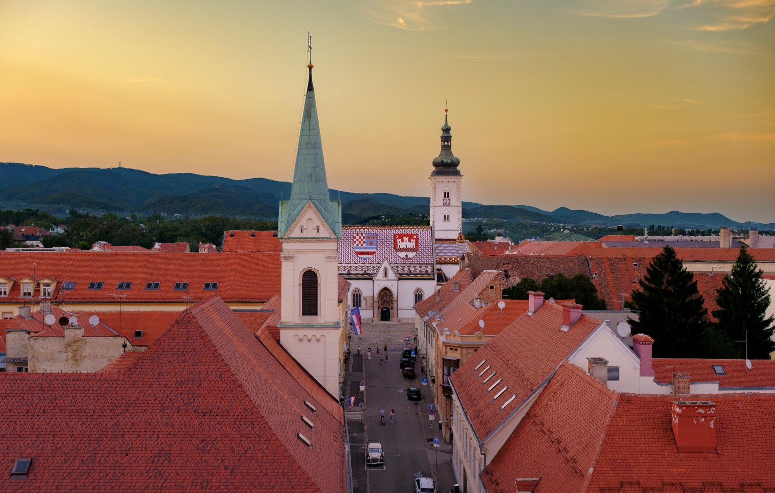 An elevated view over Zagreb's red-tiled rooftops at sunset, looking down a street towards a church with a tiled mosaic roof and twin towers.