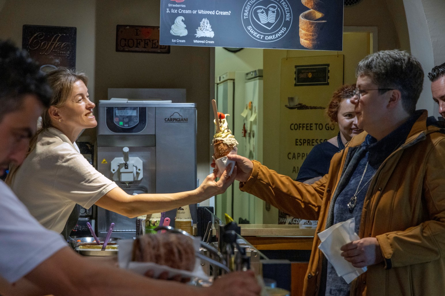 A vendor handing a customer a soft-serve ice cream cone topped with whipped cream and strawberries at a Prague trdelník shop.
