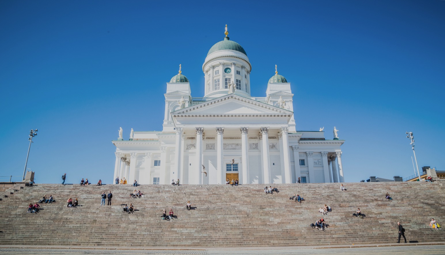 Helsinki Cathedral's white neoclassical facade and green domes, with people sitting on the steps below