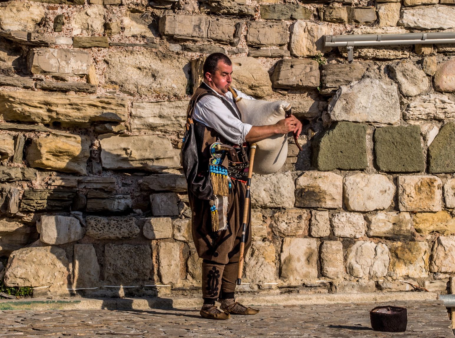 A street performer in traditional Bulgarian dress playing a gaida (bagpipe) against an old stone wall in Nessebar