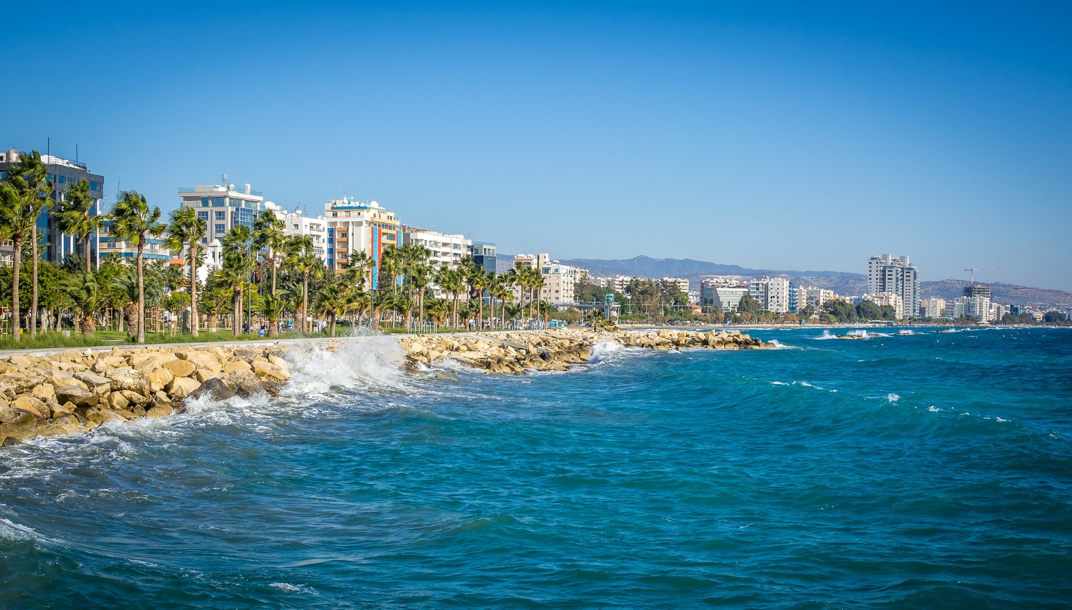 Waves breaking against a rocky breakwater along the palm-lined seafront of Limassol, Cyprus
