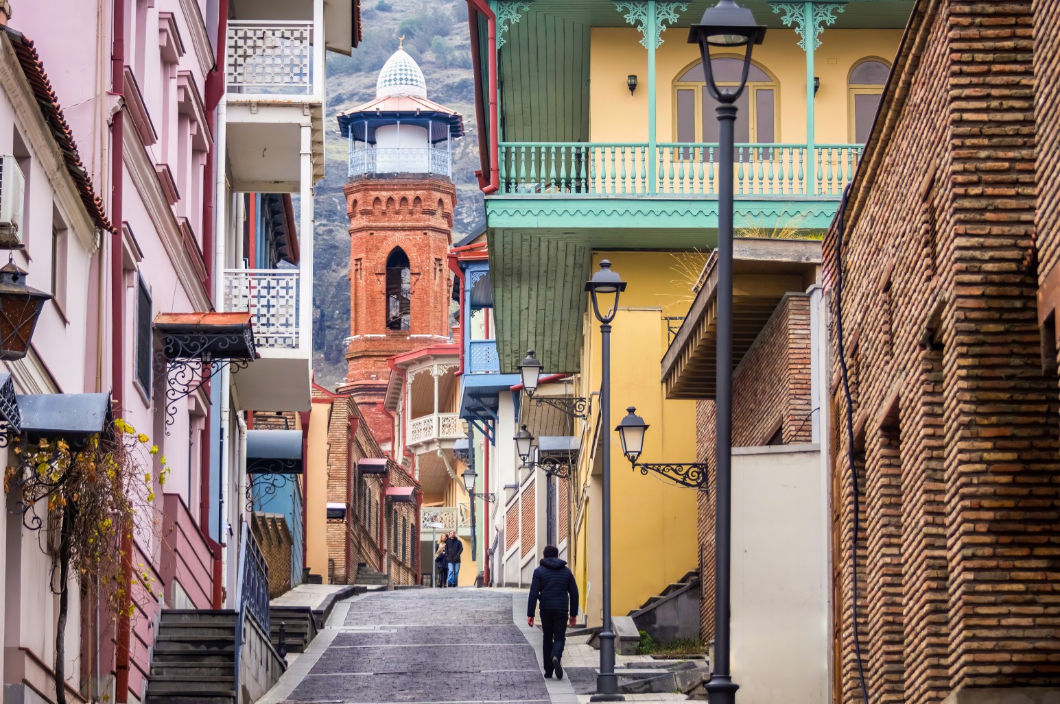 Narrow cobbled street in Old Tbilisi lined with colourful carved wooden balconies, with a brick minaret rising in the background