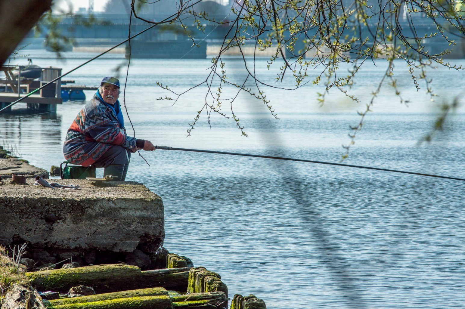 An elderly man fishing with a long rod from a concrete pier beside a calm canal, with a bridge and moored boats visible in the background.