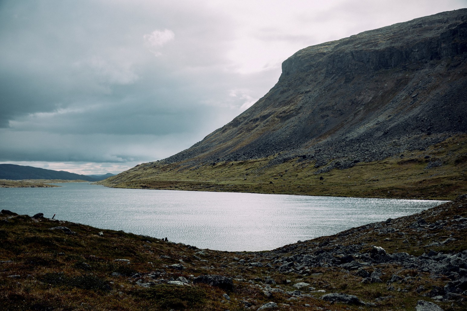 A still lake beneath a rocky, moss-covered fell under a cloudy sky in Kilpisjärvi, Finland
