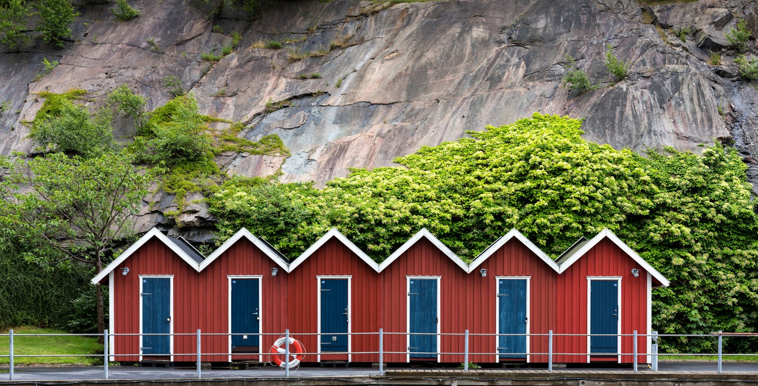 A row of red wooden boathouses with blue doors built into a rocky, tree-covered hillside in Gothenburg, Sweden