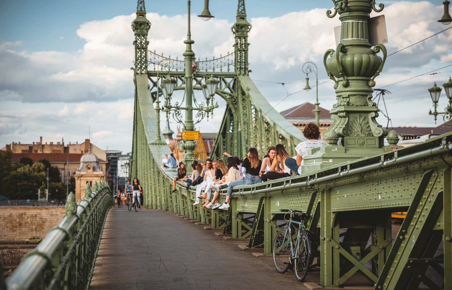 People sitting and chatting along the green ironwork of Liberty Bridge in Budapest, with a bicycle parked against the railing