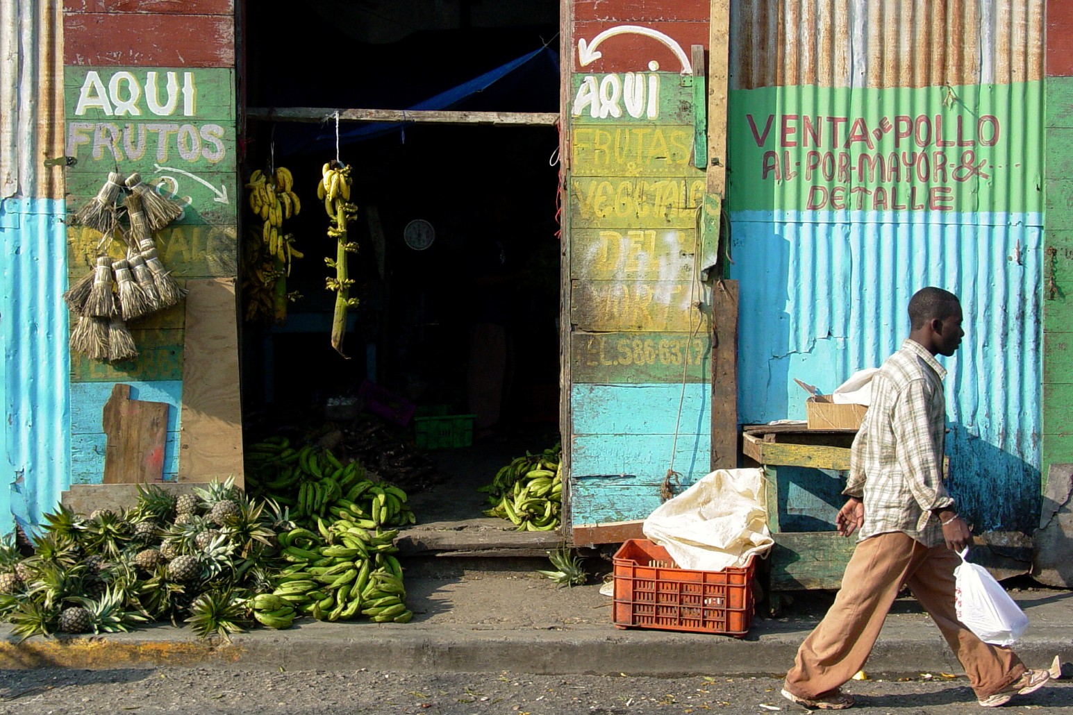 A man walking past a colourful corrugated metal shopfront in Puerto Plata, Dominican Republic, with bunches of plantains, bananas and pineapples piled outside and hand-painted Spanish signs advertising fruit, vegetables and chicken