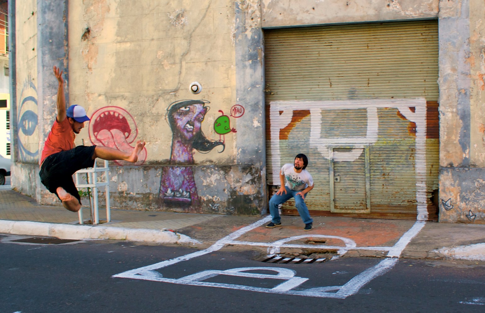 Two men playing street football beside a graffiti-covered wall in Asunción, one leaping mid-air to strike the ball while the other crouches in a goal marked out in white paint on the shutters behind him.