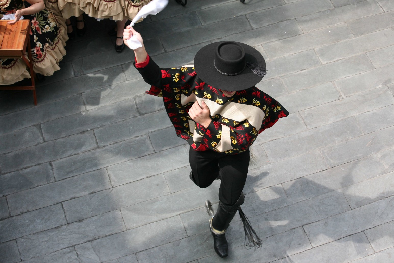 Overhead view of a cueca dancer in a black hat, patterned poncho and riding boots, waving a white handkerchief as he performs on cobblestones, with women in traditional dresses watching from the side.