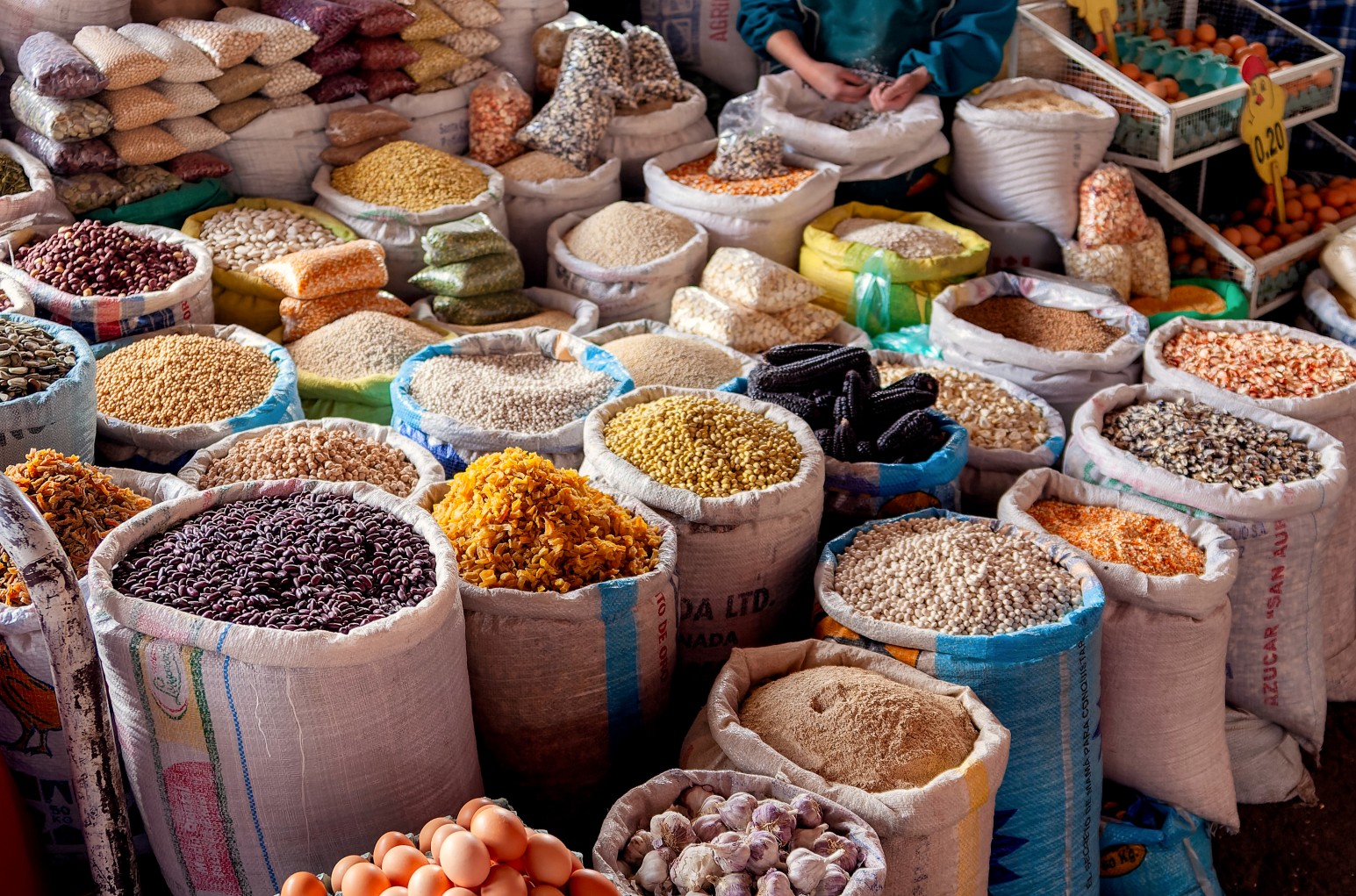 Sacks of grains, beans, and other market produce on display, with eggs and garlic in the foreground, at a market in Cusco